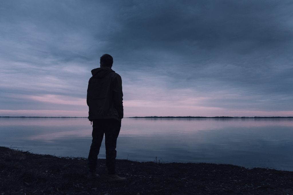 Man in dark-colored jacket standing on a lakeshore.