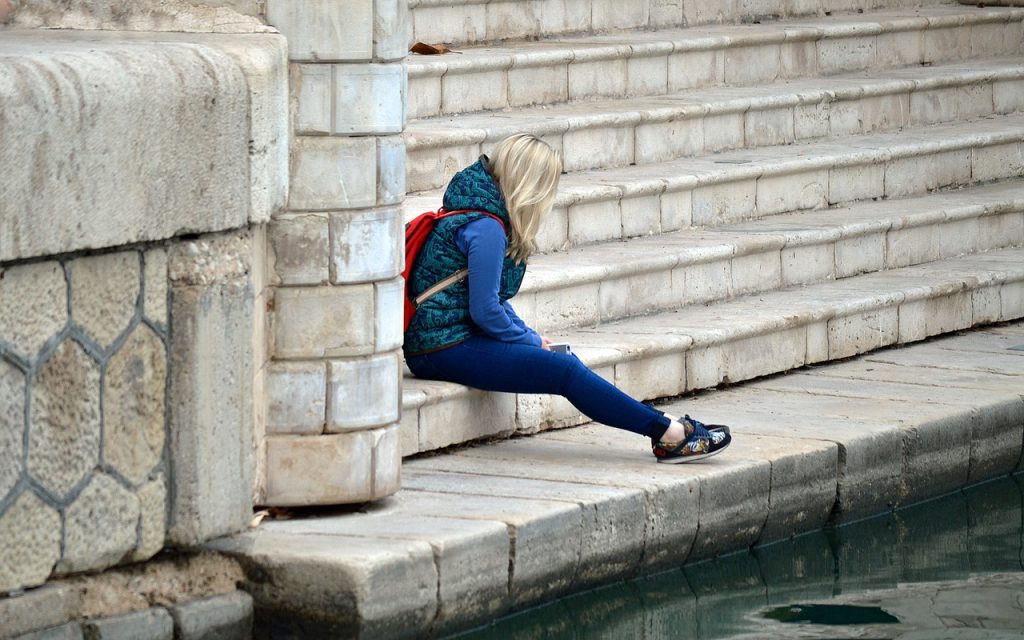 Blonde woman wearing jeans and a blue T-shirt and jacket with a red backpack sitting on a step at water's edge.