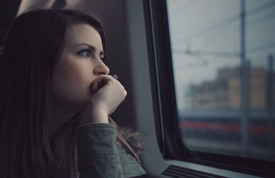 Sad-looking woman sitting with her hand on her mouth near a window.