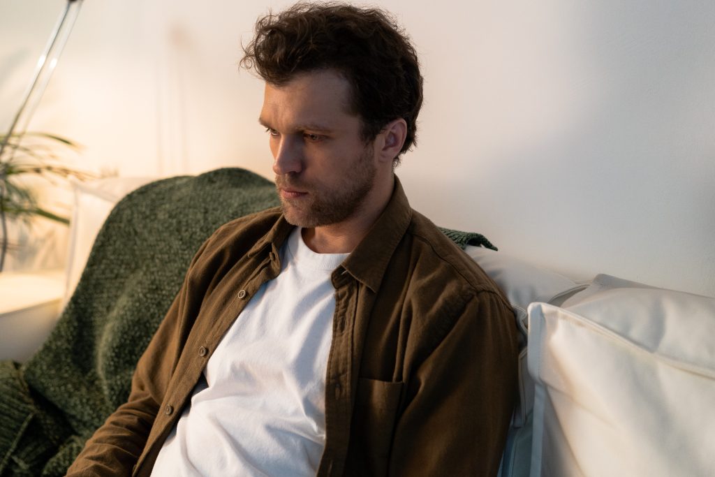 Man in white t-shirt and brown overshirt seated on a sofa looking sad.