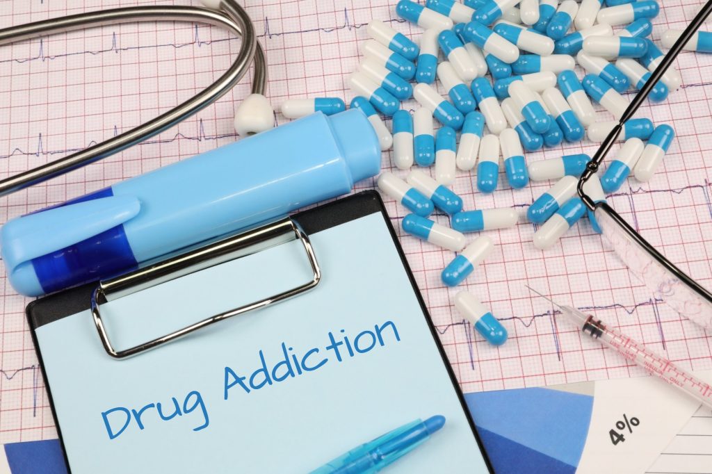 A clipboard with the header Drug Addiction along with a blue highlighting pen, a hypodermic needle, and blue and white capsules.
