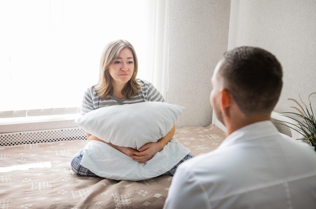 A young woman sitting on a bed with a pillow in her lap in front of a male healthcare professional in a white coat for a therapy session.
