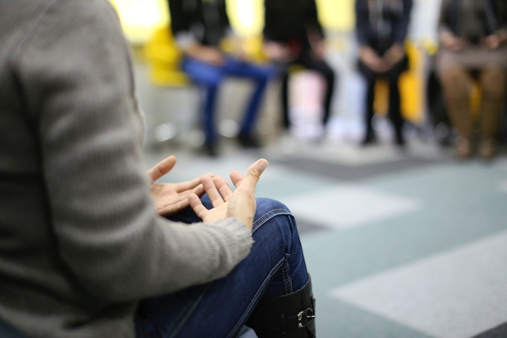 A therapist sitting with their hands in their lap in the foreground with silhouettes of four other people in the background during a group therapy session.