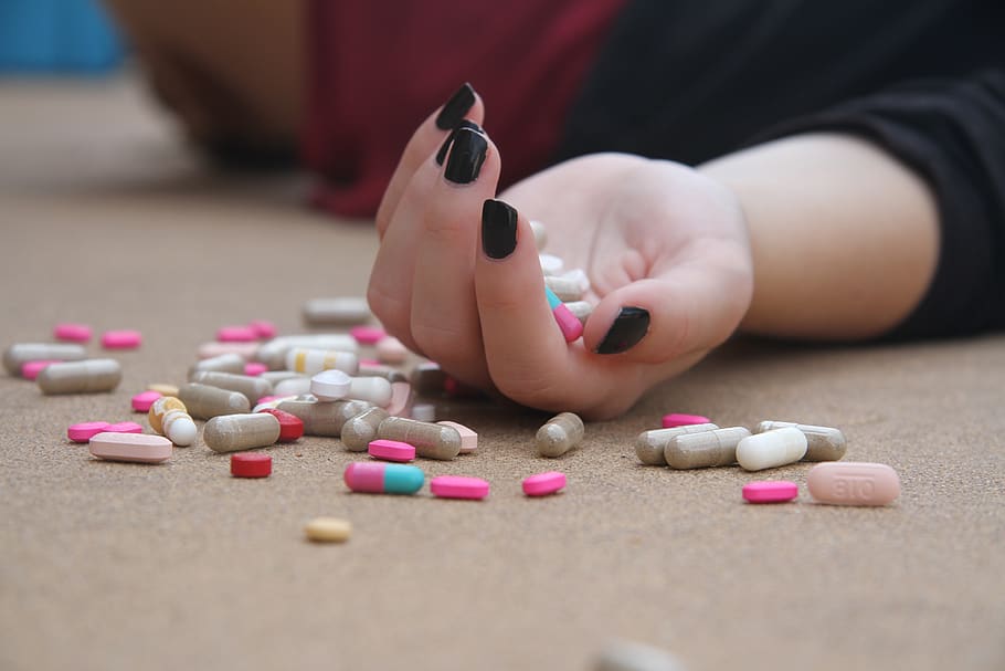 A hand with dark-colored nail polish holding several multi-colored pills and many pills scattered on the floor next to the hand. 