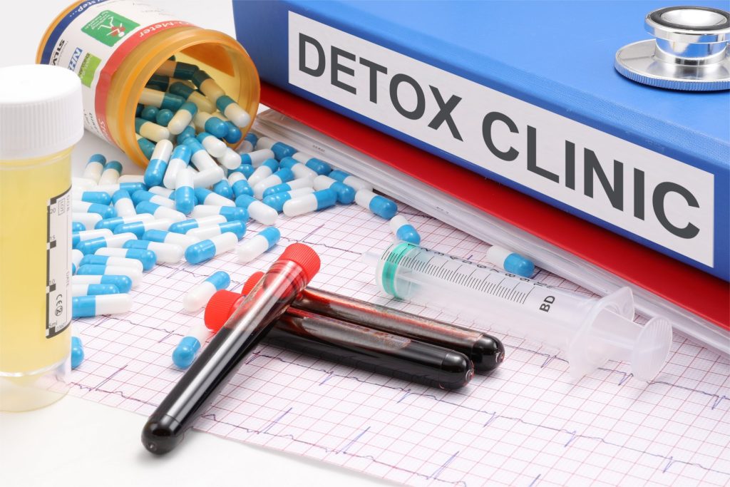 A banner that says Detox Clinic in the background with a syringe, vials of blood, and blue and white capsules spilling out of a medicine bottle in the foreground.