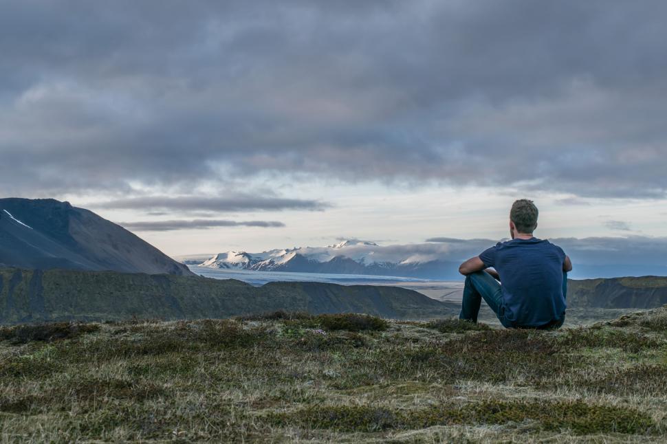 Lonely man sitting with his back to the camera in a meadow looking at a snow capped mountain range. 