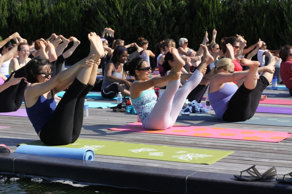 A group of women sitting on yoga mats with their legs raised as part of an outdoor yoga class.