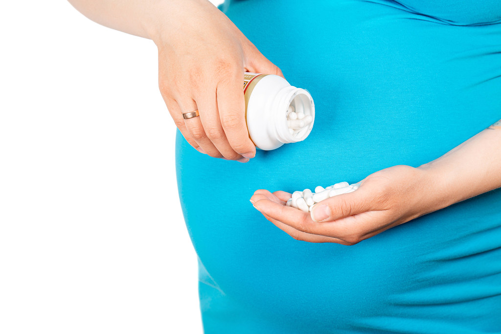 Close up of a pregnant woman's belly in a blue t-shirt with a bottle of pills in one hand and several white pills in the other hand.